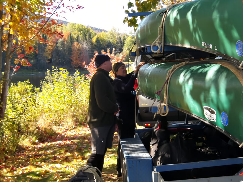 A man and a woman are standing next to a trailer carrying two green canoes. The canoes are stacked on top of each other and secured to the trailer. The man is wearing a black hat and a gray jacket, while the woman is wearing a black jacket. The trailer is parked on a dirt road next to a body of water. There are trees with fall foliage in the background.